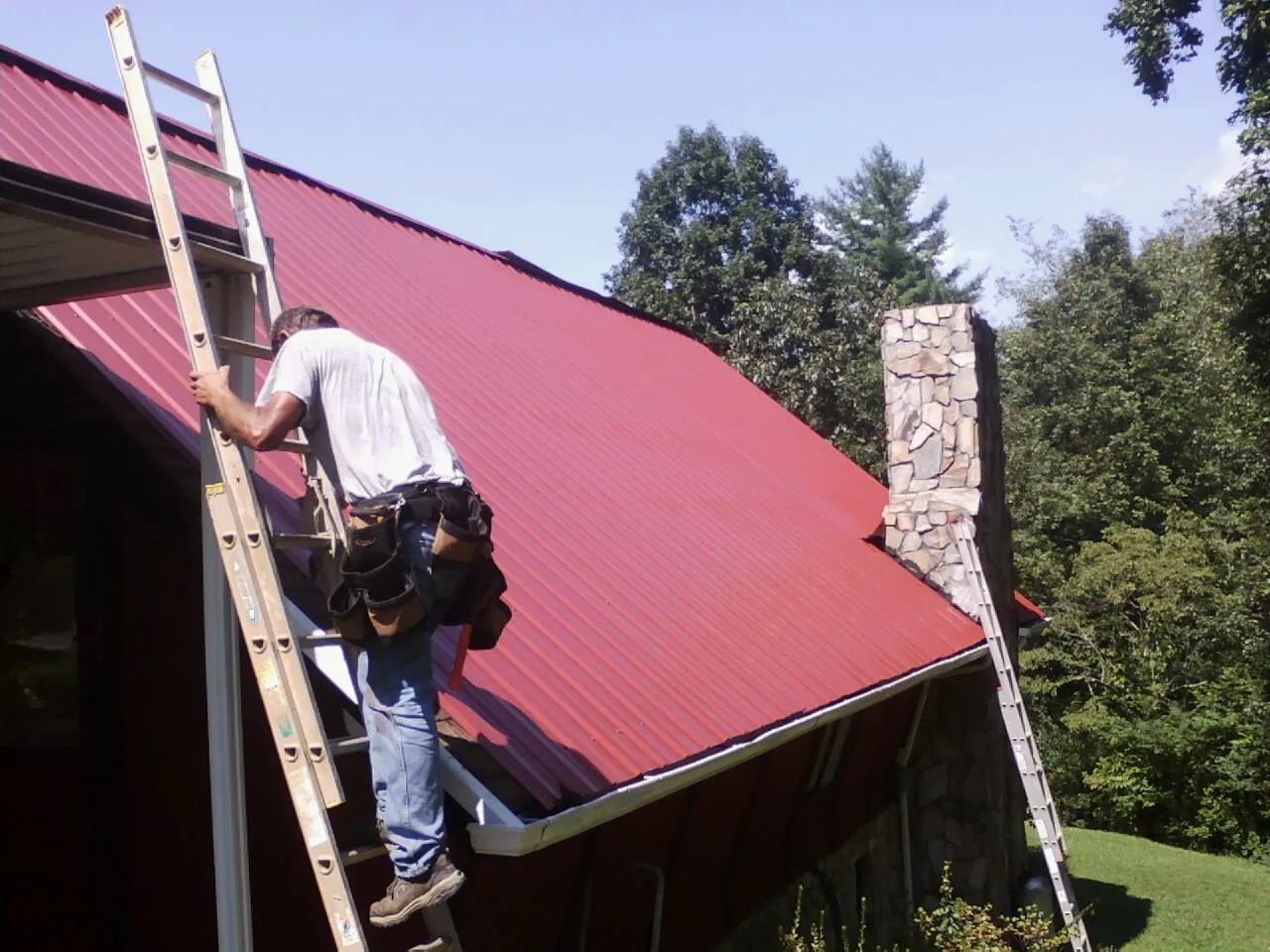 Roofer on a ladder beside a red metal roof and stone chimney — Soffit & Fascia Repair by Roofing Brothers in Spangle